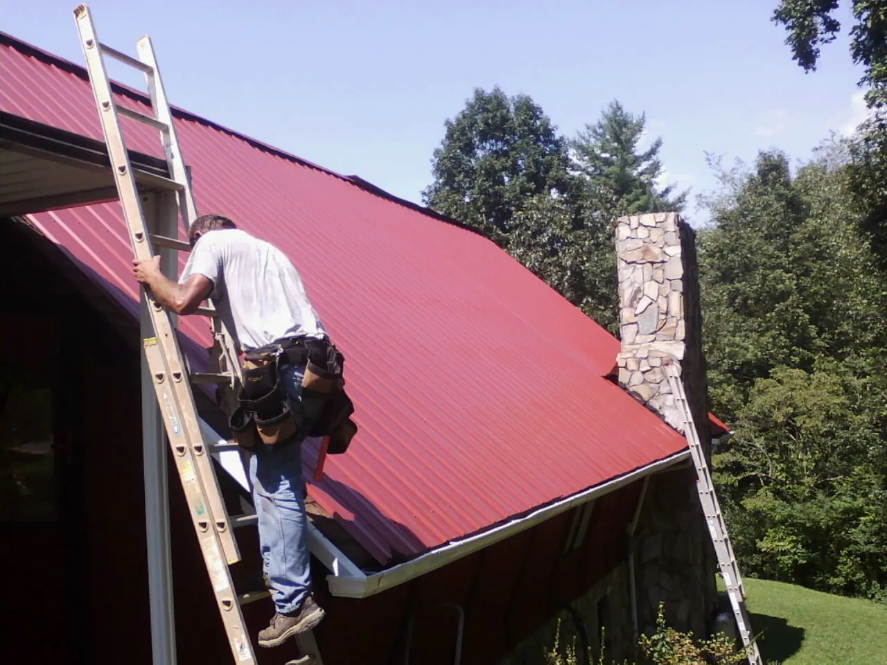 Roofer on a ladder beside a red metal roof and stone chimney — Soffit & Fascia Repair by Roofing Brothers in Arch Cape